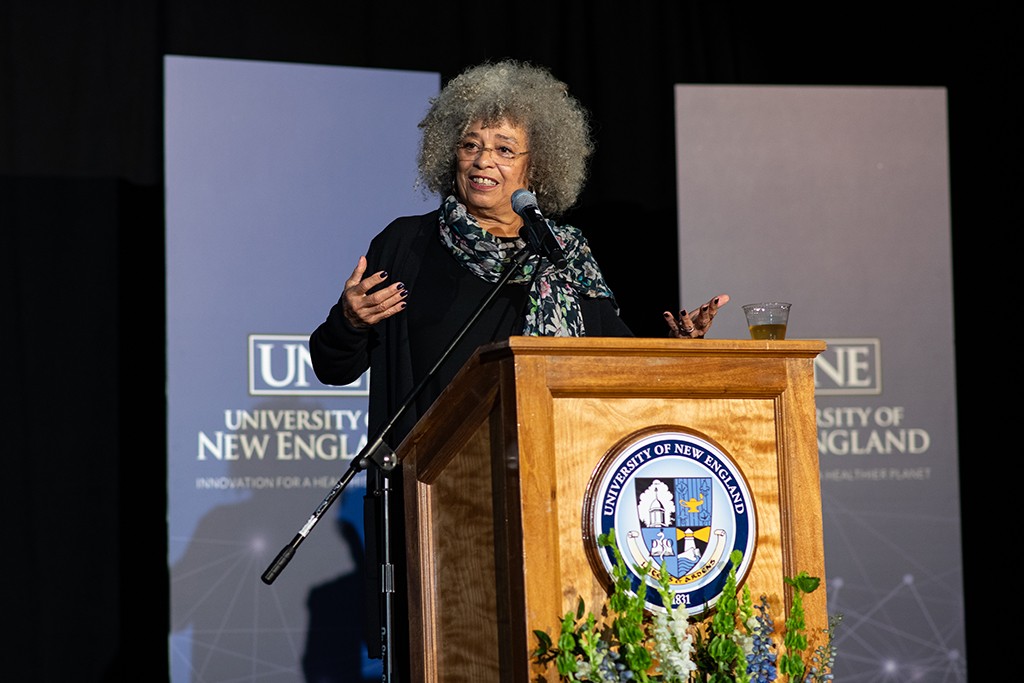 Angela Davis stands at a podium in front of a screen displaying the U N E logo
