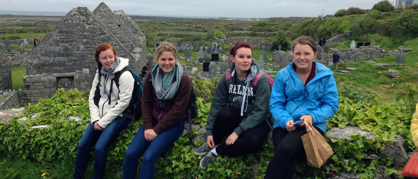 A group of U N E students sit on a stone wall at a rolling hillside cemetery in Ireland