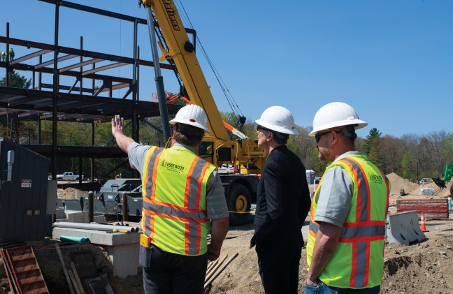 Two men stand with the U N E President as they survey the construction site for the upcoming health sciences building